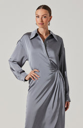 A woman is wearing a shiny, silver-gray satin dress with a classic collar, long sleeves, and a wrap-style design that gathers fabric on the right side, creating a draped effect. The dress appears elegant and smooth, with a sleek and polished appearance.